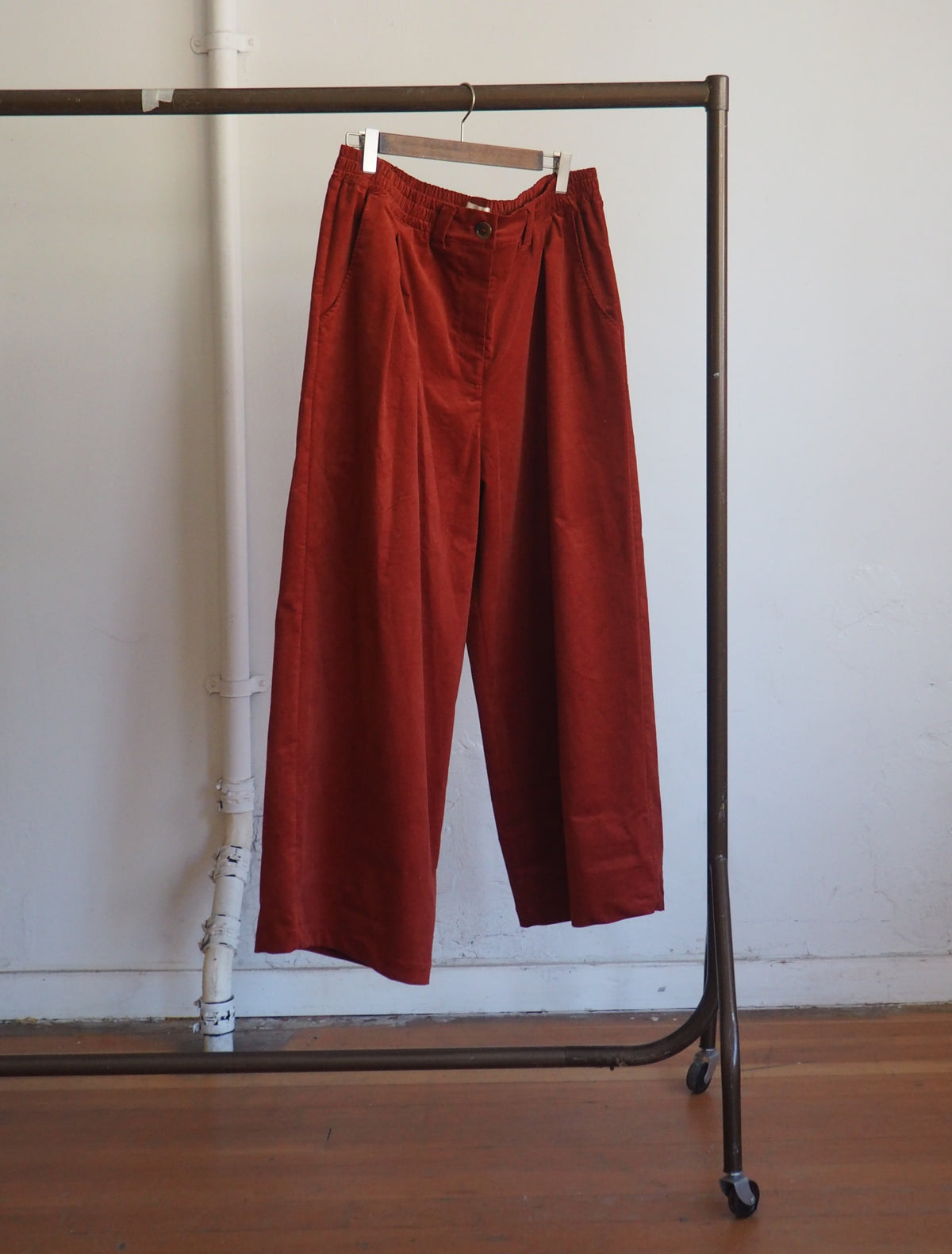 A pair of rust-orange wide-leg cropped trousers hanging on a clothing rack against a white wall with a wooden floor.