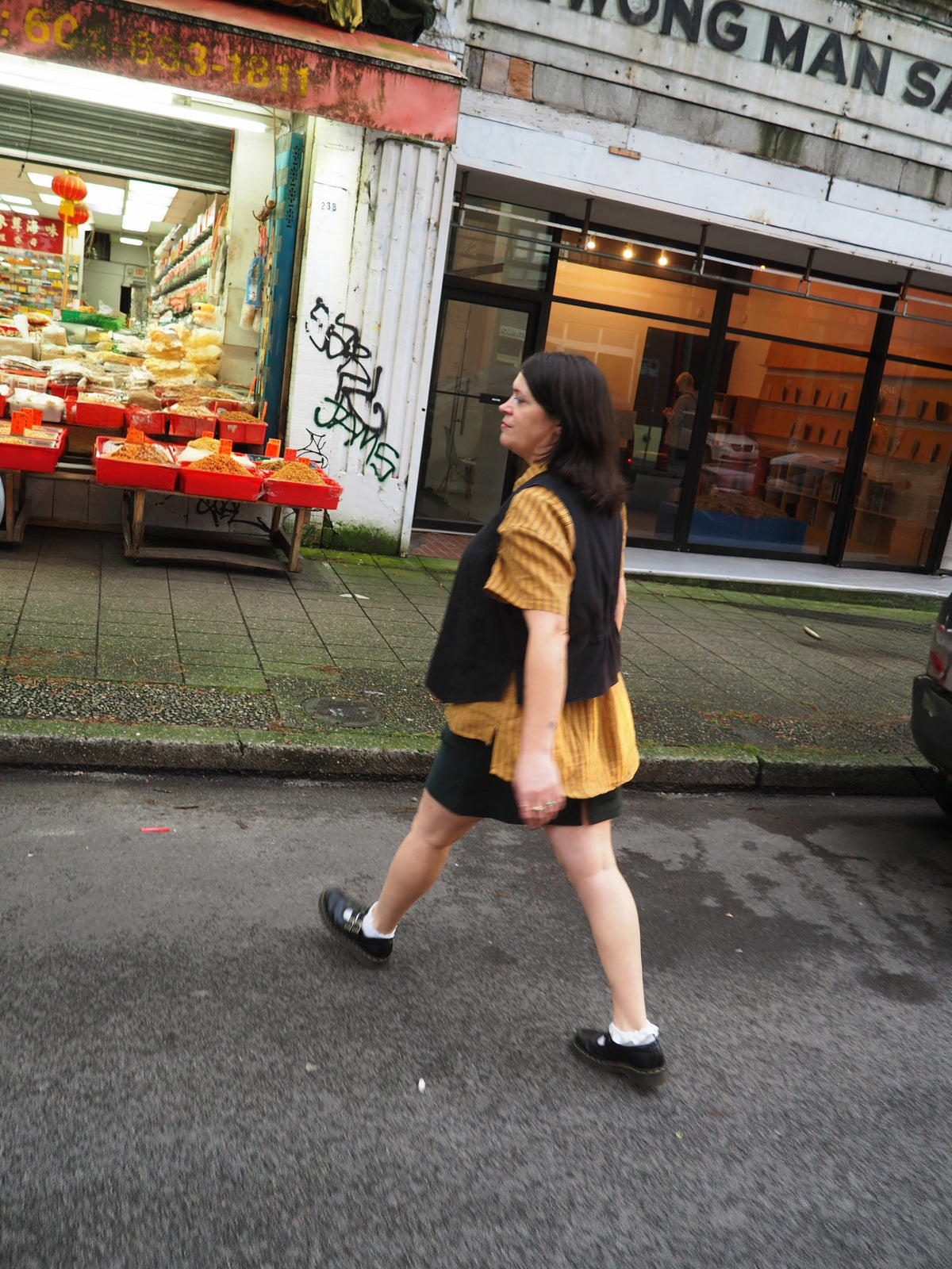 A person wearing a yellow top, black vest, and black skirt walks across a street in front of a shop.