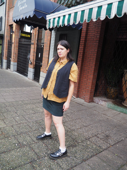A person stands on a city sidewalk wearing a yellow striped short-sleeve shirt, a dark navy vest, a dark skirt, and black Mary Jane shoes. Brick storefronts with green-and-white awnings are in the background.