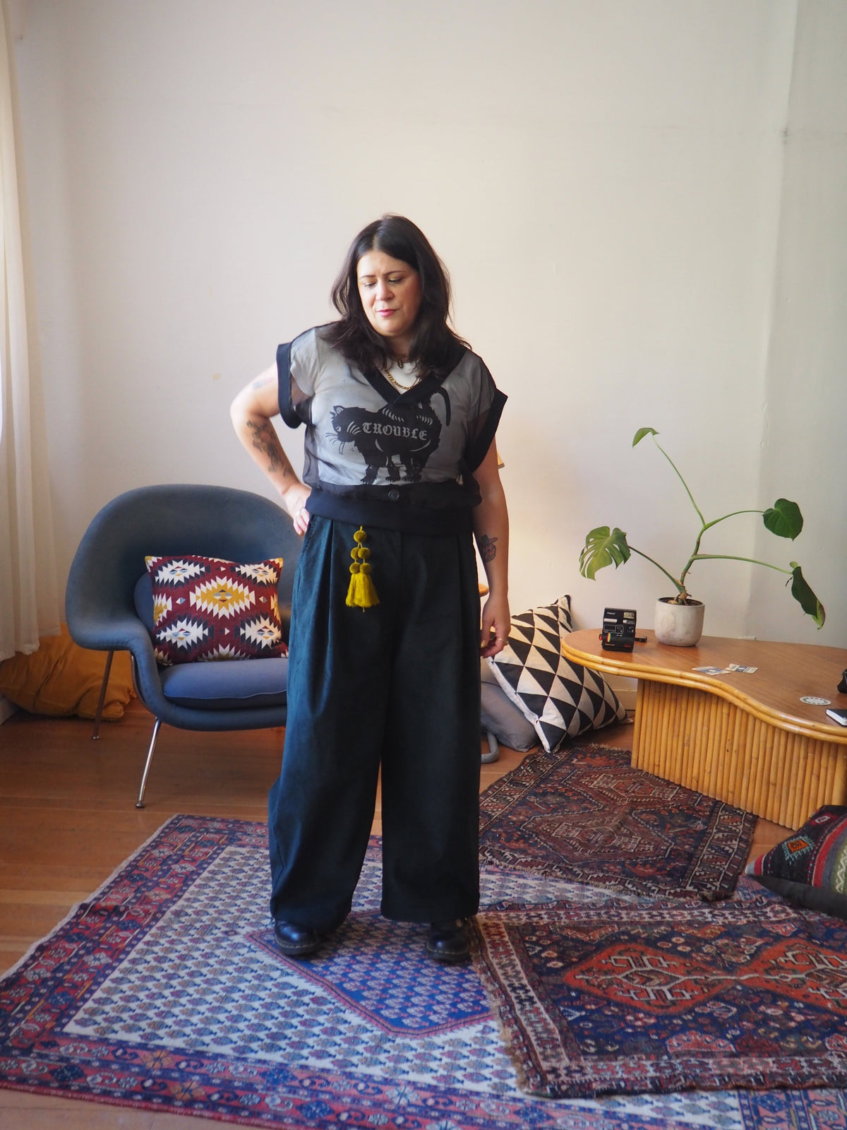 A person stands in a room wearing a black graphic t-shirt and wide-leg dark trousers with a yellow tassel belt. The room features a blue chair, a wooden side table with a plant, and patterned rugs on the floor.
