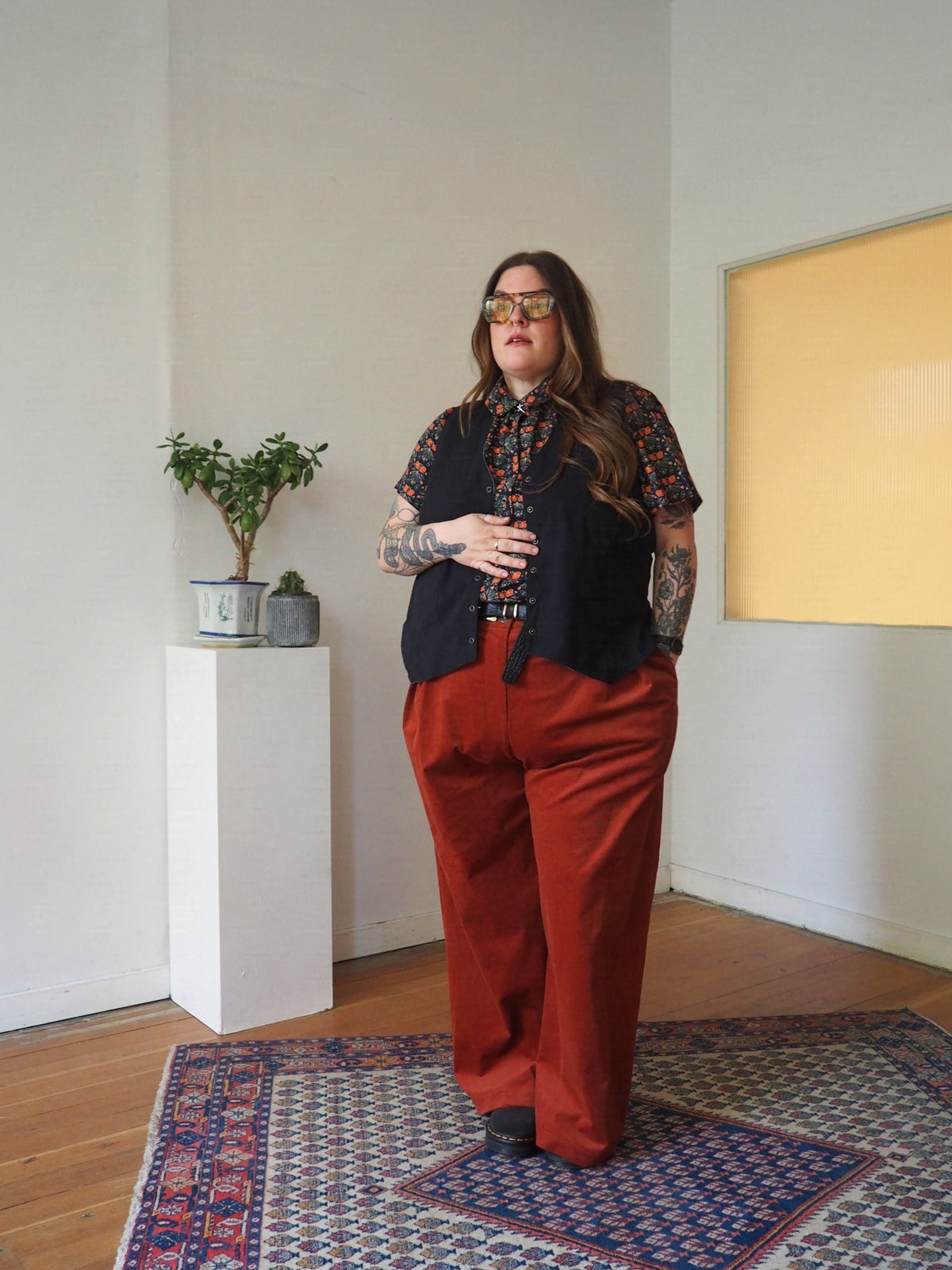 A person stands indoors wearing a black vest over a short-sleeve patterned shirt and rust-red wide-leg pants, with gold-framed sunglasses. The room has white walls, a wooden floor, a patterned rug, and a small plant on a white pedestal.