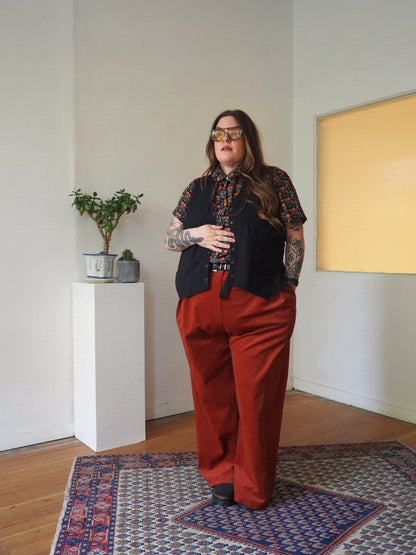 A person stands indoors wearing a black vest over a short-sleeve patterned shirt and rust-red wide-leg pants, with gold-framed sunglasses. The room has white walls, a wooden floor, a patterned rug, and a small plant on a white pedestal.