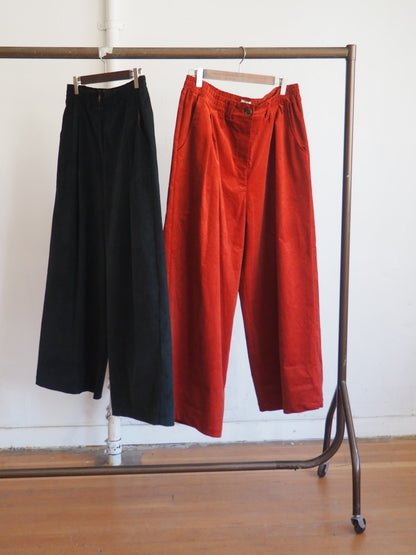 Two pairs of wide-leg cropped trousers hanging on a clothing rack against a white wall and wood floor. The left pair is dark green, and the right pair is a warm rust red-orange.