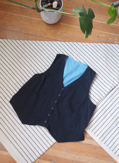 A sleeveless black vest with a light blue inner lining and a buttoned front, laid flat on a cream and black striped rug over a wooden floor.