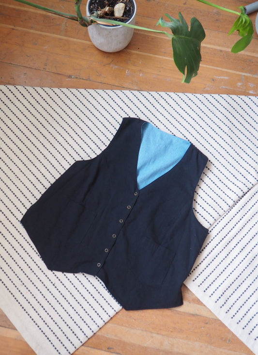 A sleeveless black vest with a light blue inner lining and a buttoned front, laid flat on a cream and black striped rug over a wooden floor.