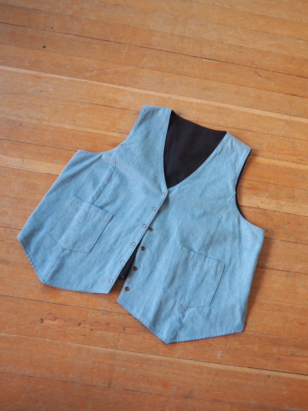 A light blue denim vest with a black lining, laid flat on a wooden floor.