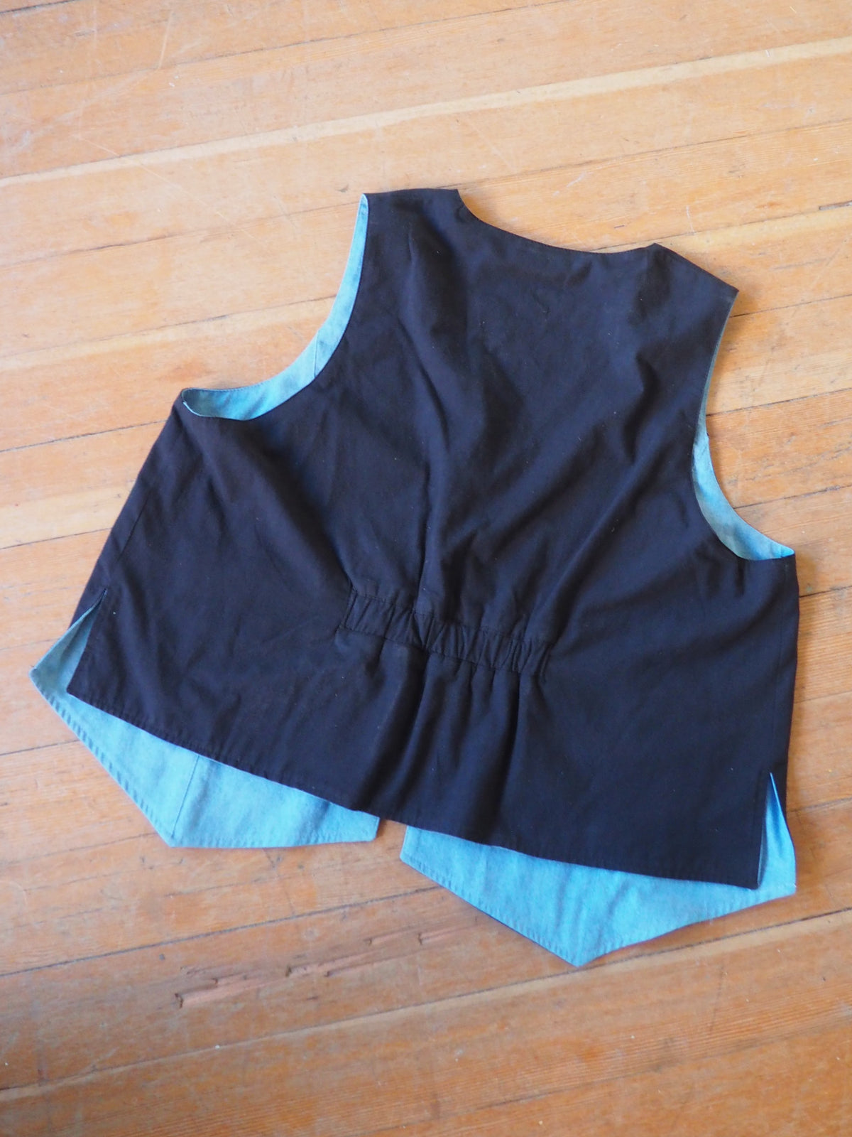 A sleeveless black vest with a light blue lining, laid flat on a wooden floor.