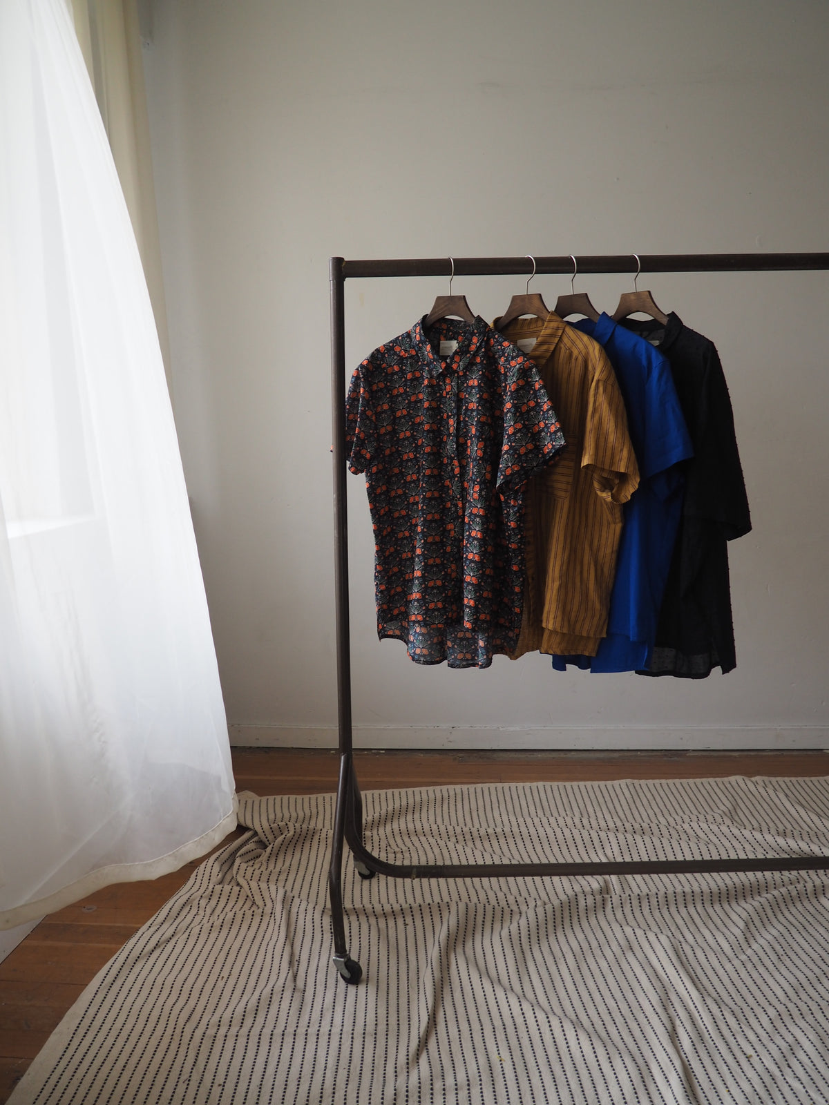 A stack of folded clothing in multiple colors, including black, blue, brown, and a patterned shirt, hanging on a black clothing rack positioned next to a bed with striped bedding.