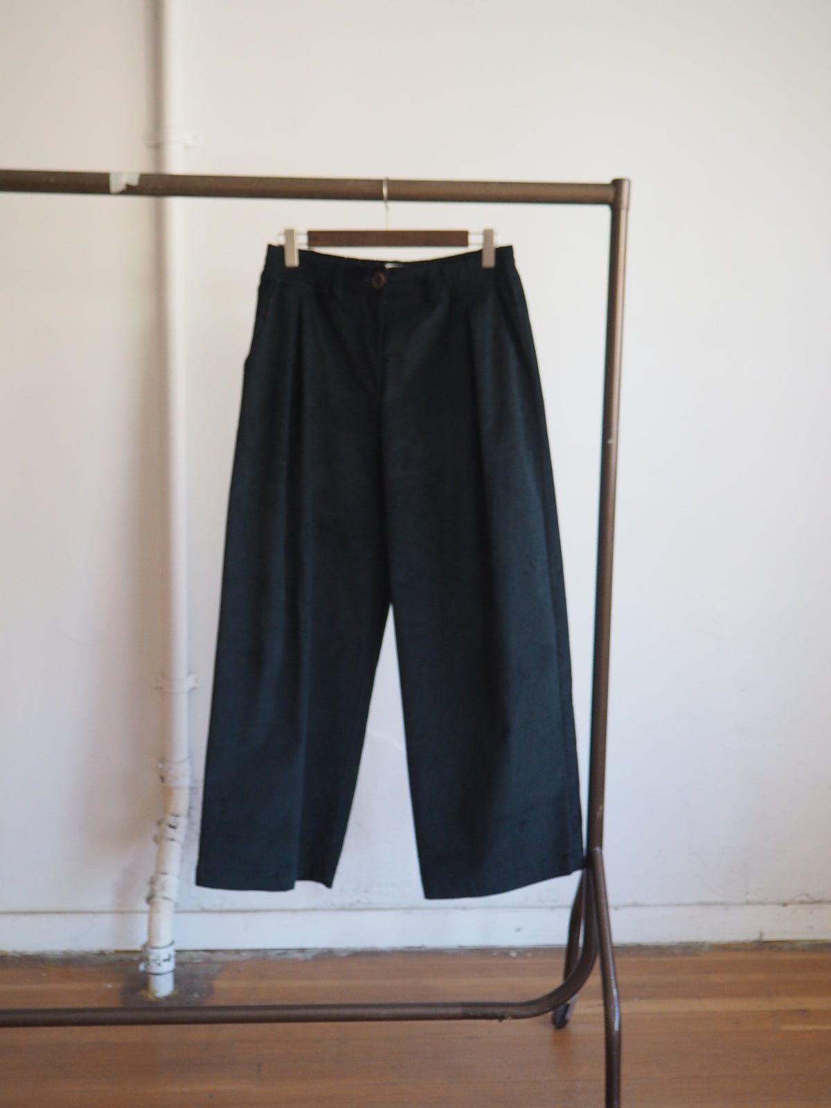 Dark Green wide-leg cropped trousers hanging on a wooden hanger from a metal clothing rack against a white wall and wood floor.