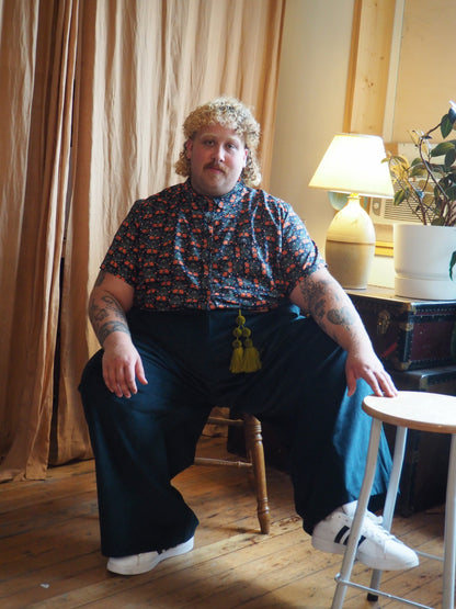 A person with curly blonde hair and a mustache sits on a wooden chair in a room with beige curtains and a wooden floor. The person is wearing a short-sleeve button-up shirt with a red, blue, and white floral pattern, dark wide-leg trousers, and white sneakers.