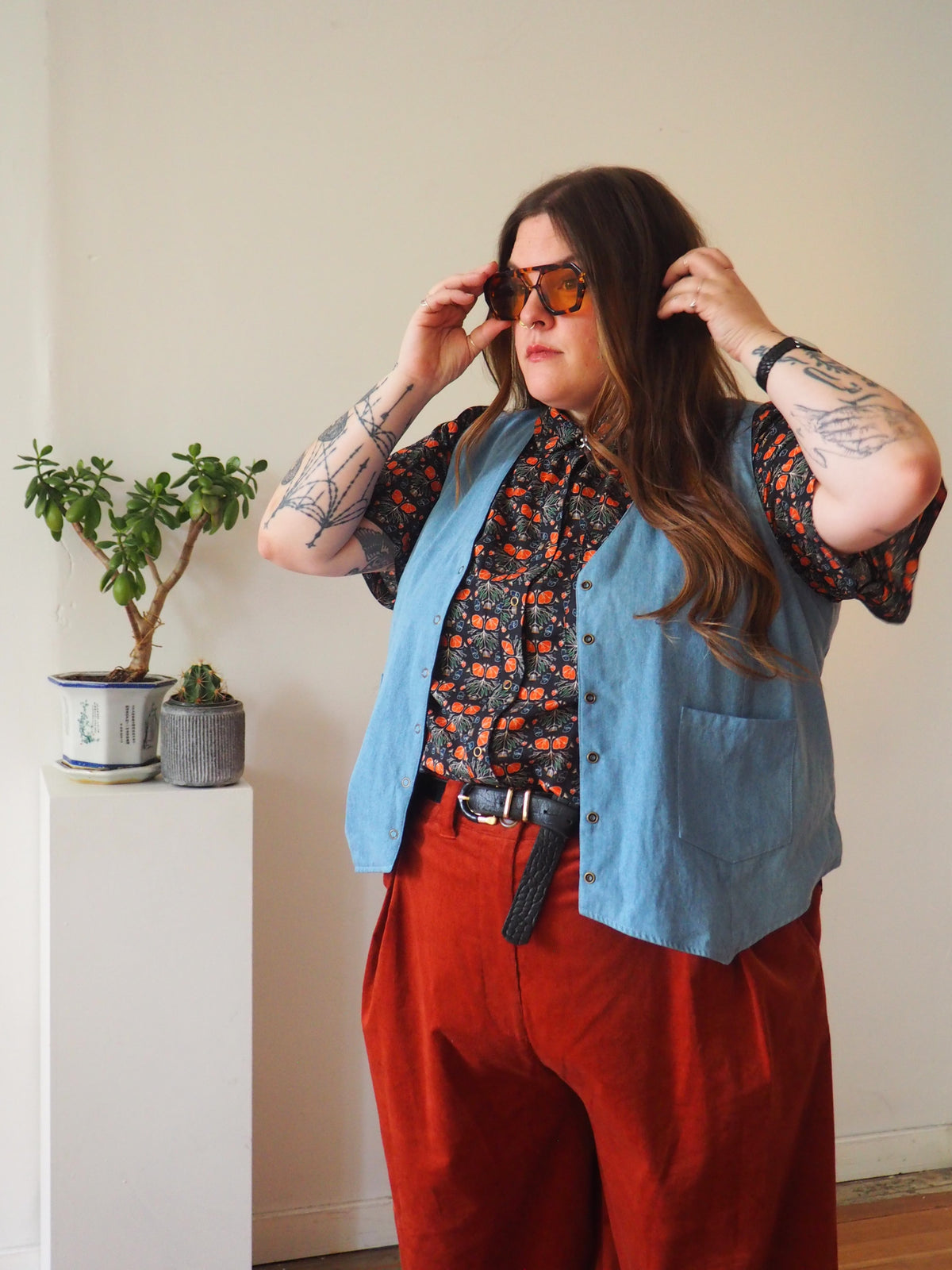A person wearing a light blue linen vest over a short-sleeve floral shirt, paired with rust-orange wide-leg trousers and tortoiseshell sunglasses, standing in a minimalist room next to a potted plant.
