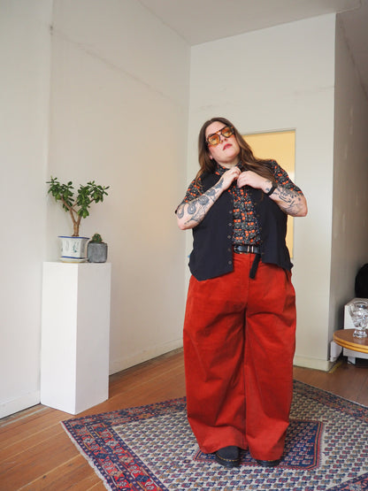 A person wearing orange-tinted sunglasses, a black vest over a colorful patterned shirt, and bright red wide-leg pants stands on a patterned rug in a minimalist room with white walls and wooden floors.