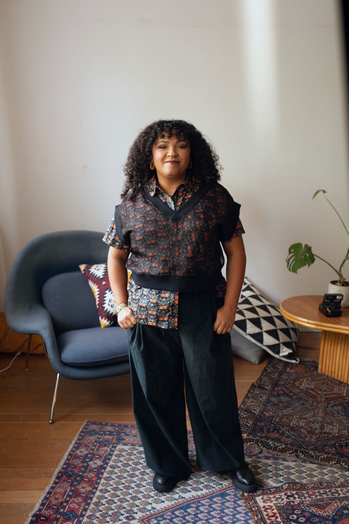 A person stands in a styled room wearing a short-sleeve button-up shirt with a dark floral pattern and wide-leg dark trousers.