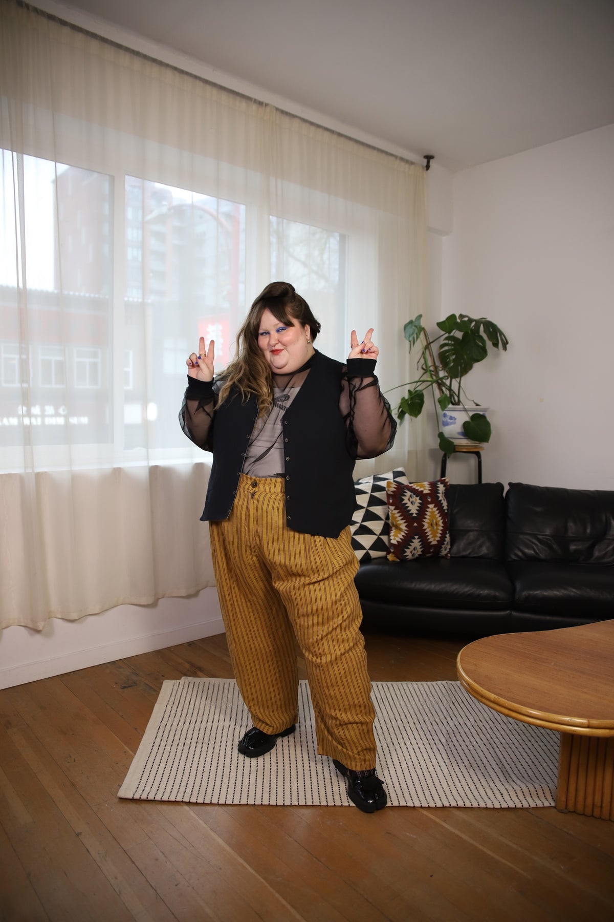A person stands in a living room wearing a black vest over a sheer long-sleeve top, a gray shirt, and mustard yellow pinstriped corduroy pants. She is posing with her hands raised in front of a window with sheer curtains, with a black leather sofa and a wooden coffee table in the background.