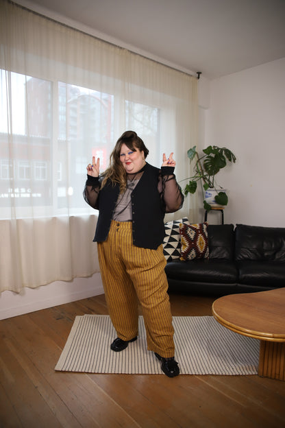 A person stands in a living room wearing a black vest over a sheer long-sleeve top, a gray shirt, and mustard yellow pinstriped corduroy pants. She is posing with her hands raised in front of a window with sheer curtains, with a black leather sofa and a wooden coffee table in the background.