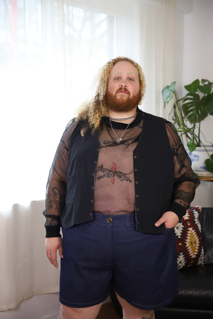A person standing indoors wearing a sheer black long-sleeve button-front overshirt over a brown graphic T-shirt, dark navy shorts, and layered silver necklaces. The background shows a bright window with white curtains, a potted plant, and part of a patterned chair.