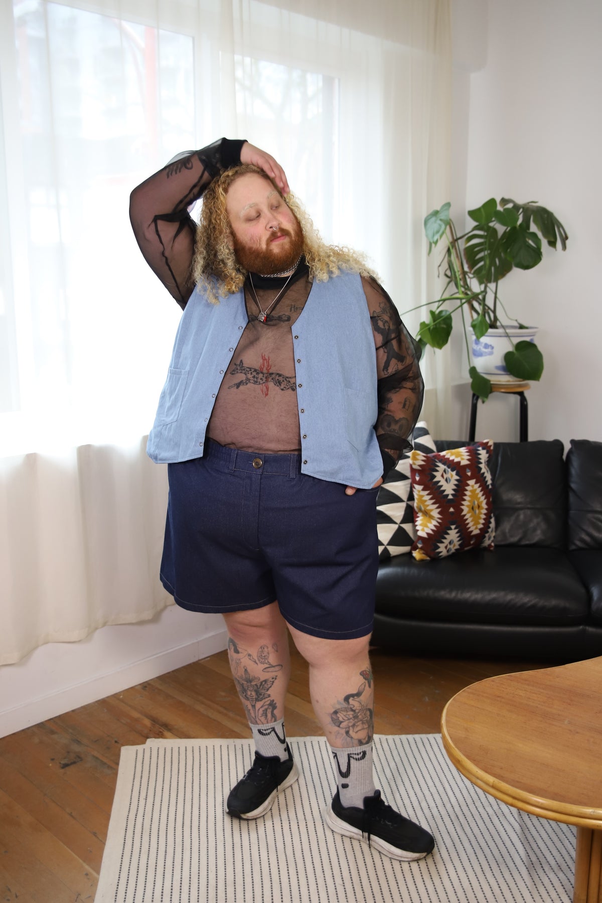 A person modeling a light blue denim vest over a sheer black long-sleeve top, paired with navy shorts, black sneakers, and patterned socks in a bright living room setting.