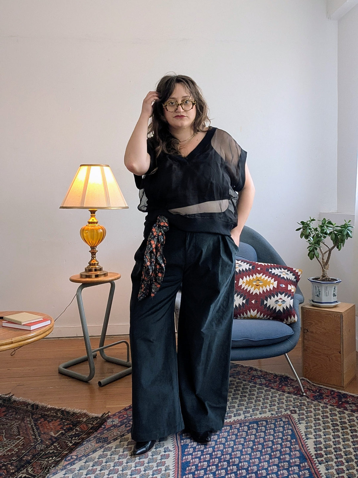 A person wearing a black sheer sleeveless top and matching wide-leg trousers with a patterned fabric tie, standing in a styled room with a lamp, chair, and rug.