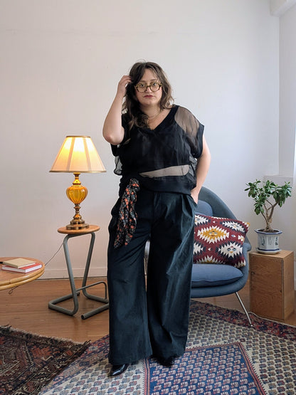 A person wearing a black sheer sleeveless top and matching wide-leg trousers with a patterned fabric tie, standing in a styled room with a lamp, chair, and rug.