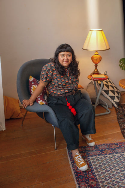 A person sits in a gray lounge chair in a cozy room with wooden floors, wearing a short-sleeve patterned top, dark wide-leg pants, and tan sneakers with red pom-pom accents. A lamp, side table, and patterned rug are visible in the background.