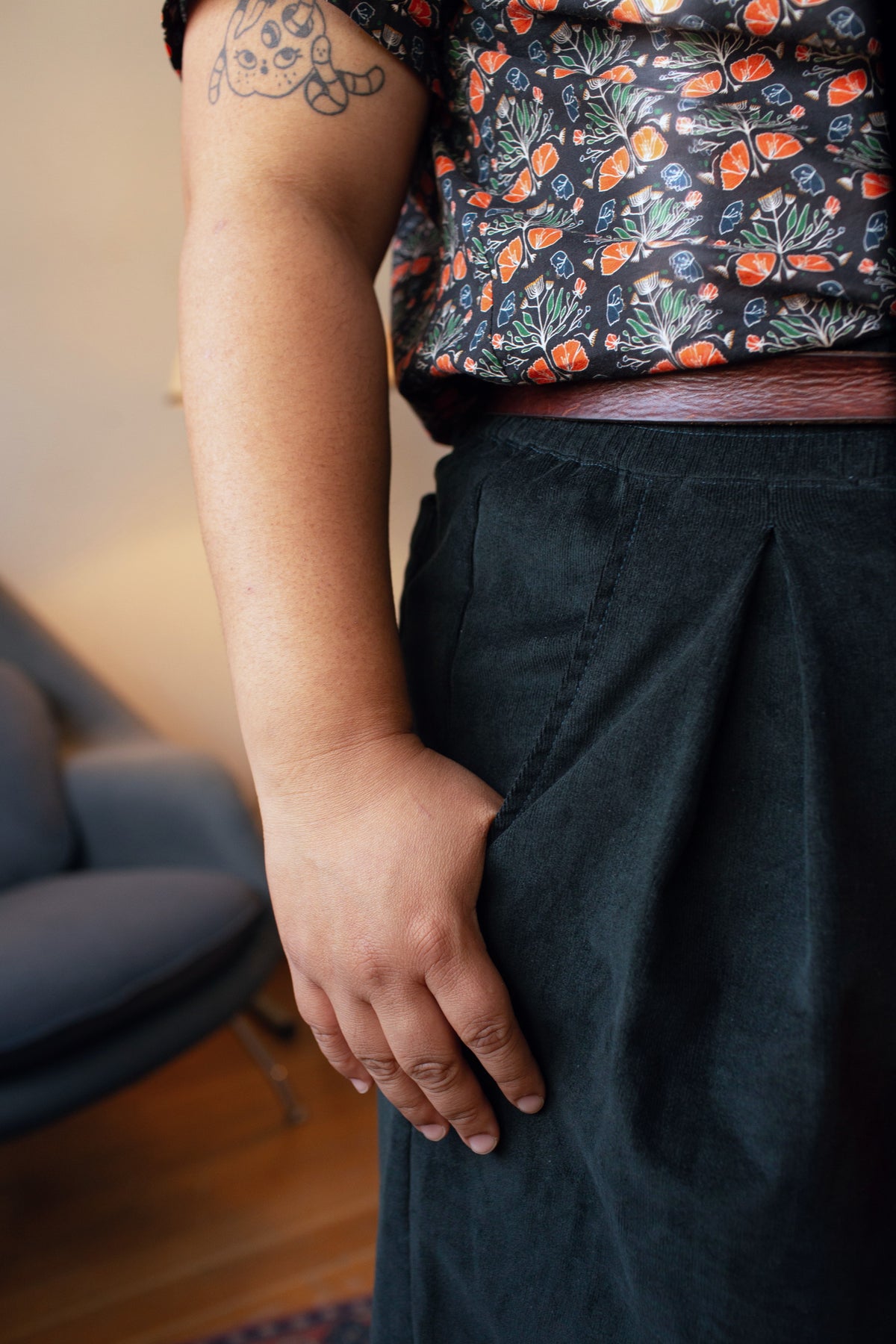 A close-up lifestyle shot of a person wearing dark charcoal gray pleated corduroy trousers and a patterned floral top, with their hand resting in the pocket of the trousers.