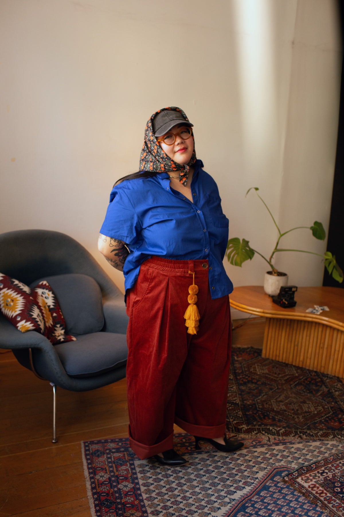 A person stands in a styled indoor room wearing a bright blue short-sleeve button-up shirt, loose dark red wide-leg pants, a patterned headscarf, and glasses. A blue chair, wooden side table, plant, and patterned rug are visible in the room.