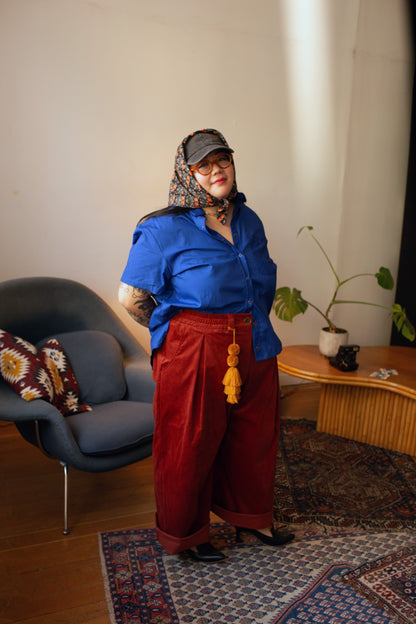 A person stands in a styled indoor room wearing a bright blue short-sleeve button-up shirt, loose dark red wide-leg pants, a patterned headscarf, and glasses. A blue chair, wooden side table, plant, and patterned rug are visible in the room.