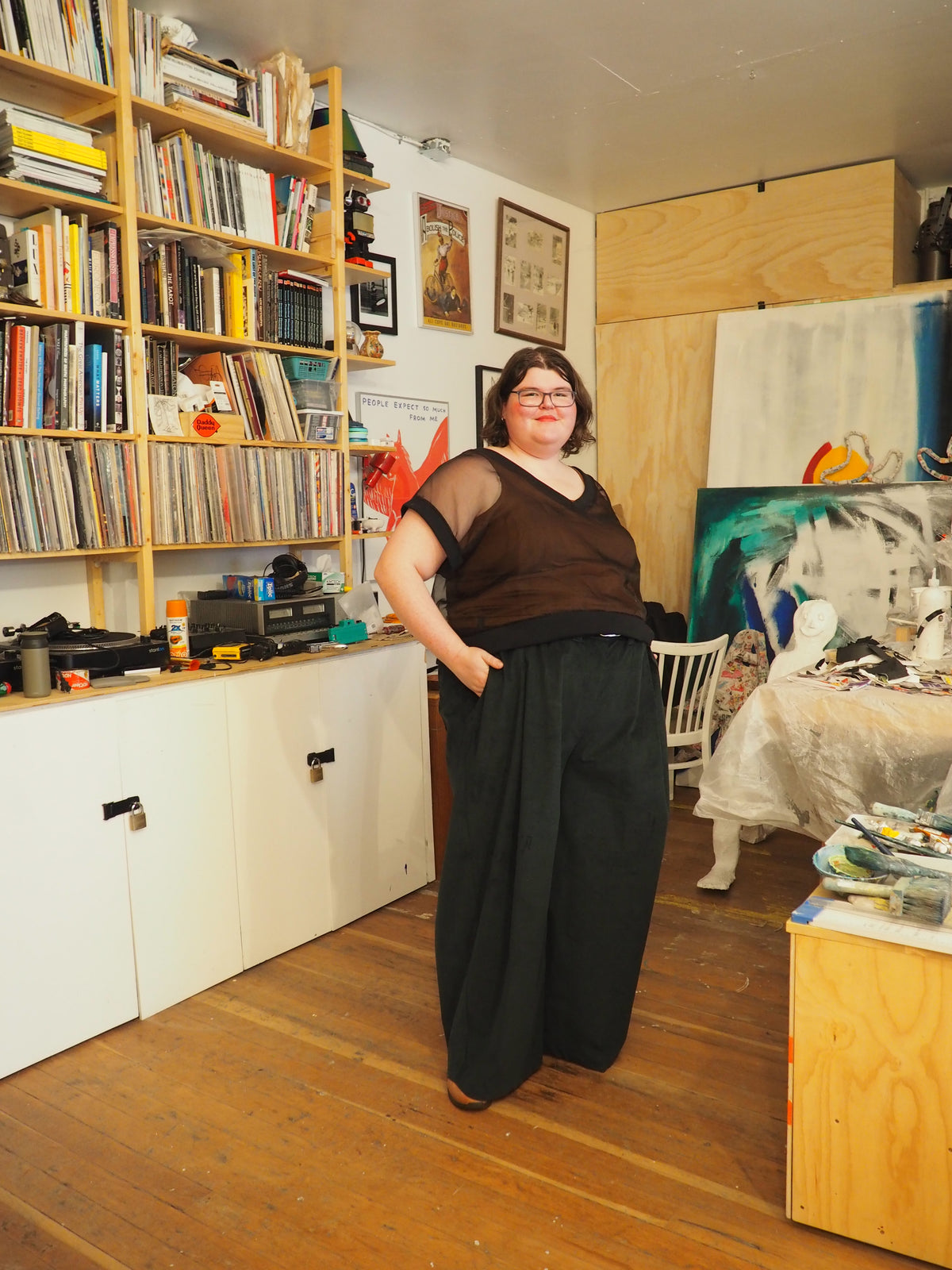 A person standing in a cluttered art studio wearing a sheer brown top and wide-leg black pants, with bookshelves and paintings visible in the background.
