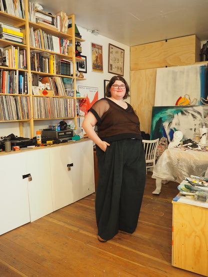 A person standing in a cluttered art studio wearing a sheer brown top and wide-leg black pants, with bookshelves and paintings visible in the background.