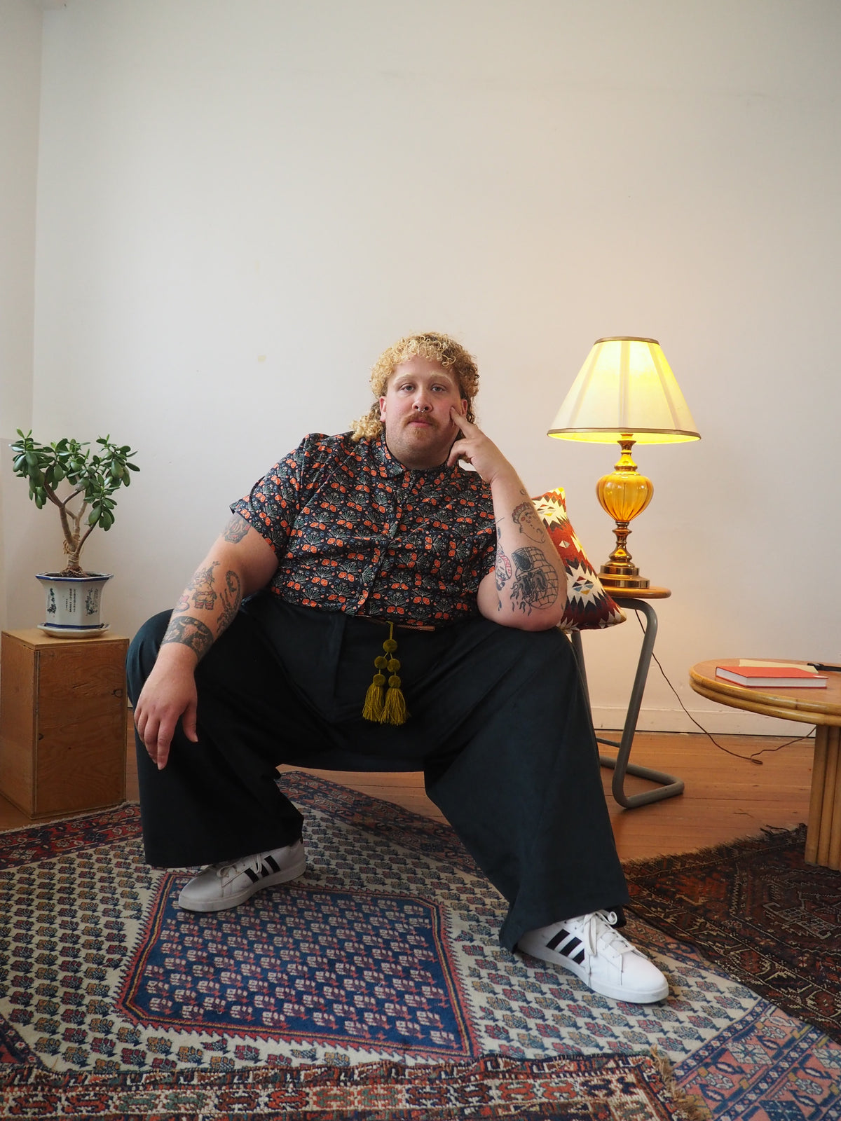 A person wearing a short-sleeve button-up shirt with a red, blue, and yellow floral pattern, black wide-leg trousers, and white sneakers, sitting on a patterned rug in a room with a lamp and plant.